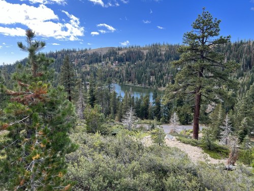 A scenic view of a lake surrounded by lush green trees and mountains under a blue sky with scattered clouds.