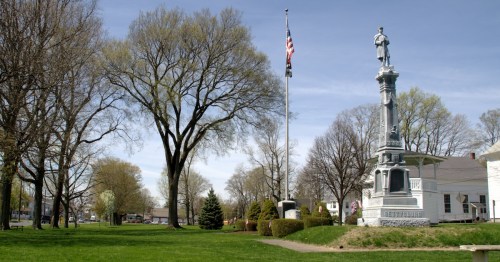 A park scene featuring a soldier monument, American flag, and trees under a clear blue sky.