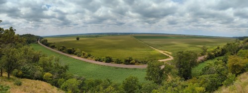 A panoramic view of lush green fields and a winding road under a cloudy sky.