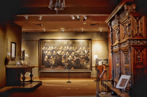 A museum interior featuring a large painting of a group portrait and ornate wooden furniture.