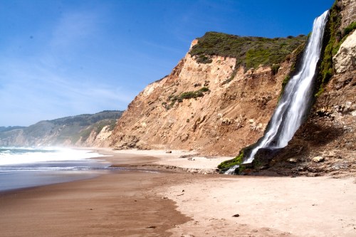 A scenic beach with a waterfall cascading down a rocky cliff under a clear blue sky.