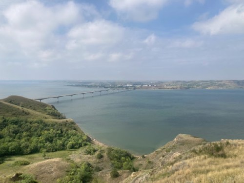 A scenic view of a bridge spanning a river, surrounded by green hills and a cloudy sky.