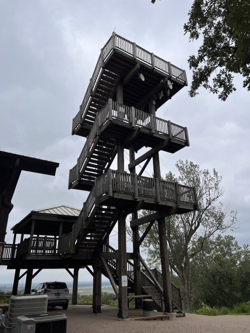 A tall wooden observation tower with multiple levels, surrounded by trees and a cloudy sky.