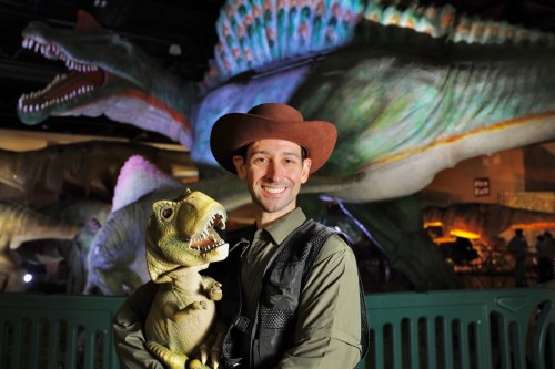 A man in a cowboy hat holds a dinosaur toy, smiling in front of large dinosaur models in a themed exhibit.
