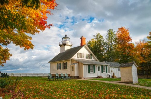 A white lighthouse and house surrounded by colorful autumn trees under a cloudy sky.