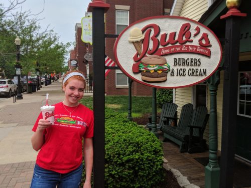 A young woman in a red shirt stands in front of Bub's Burgers & Ice Cream, holding a milkshake.