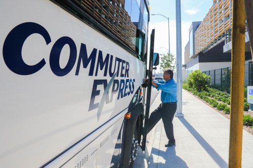 A man in a blue shirt boards a Commuter Express bus on a sunny day, with modern buildings in the background.