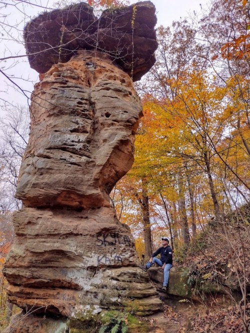 A person sits beside a tall rock formation in a forest with autumn foliage, showcasing vibrant orange and yellow leaves.