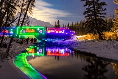 A brightly lit train decorated with colorful lights reflects in a snowy river, surrounded by trees and mountains at night.