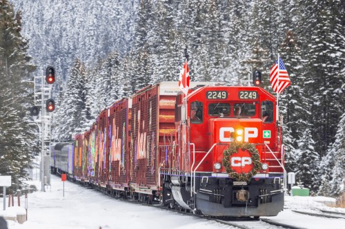 A red train with festive decorations travels through a snowy landscape, flanked by trees and signal lights.