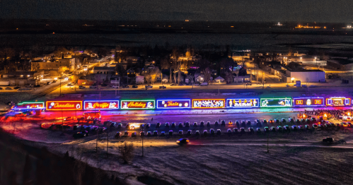 A colorful holiday light display illuminates a snowy landscape at night, with a festive train and nearby town.