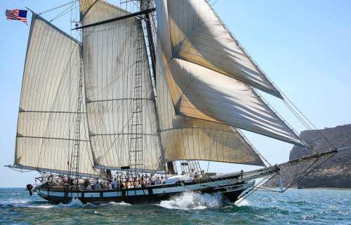A tall ship with sails raised navigates through blue waters, with a rocky coastline in the background.