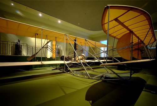 A vintage aircraft model displayed indoors, featuring a wooden frame and large fabric wings.