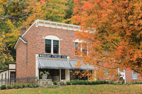 A brick building with "Wright Cycle Co." sign, surrounded by vibrant autumn foliage and a striped awning.