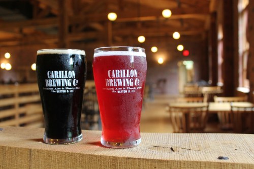 Two glasses of beer, one dark and one red, sit on a wooden ledge in a rustic brewery setting.