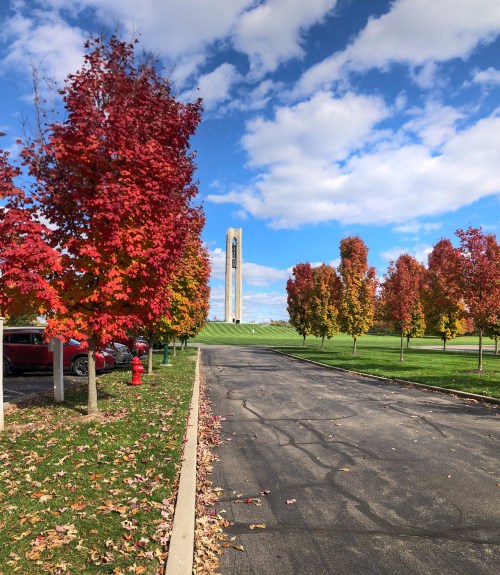 A tree-lined path with vibrant red foliage leads to a tall tower under a blue sky with scattered clouds.