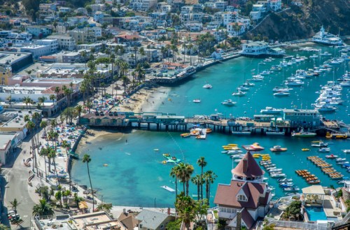 Aerial view of a coastal town with a marina, boats, sandy beach, and palm trees under a clear blue sky.