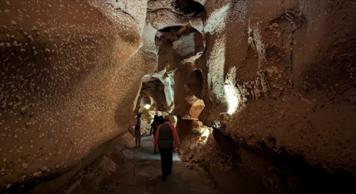 A dimly lit cave with textured walls, featuring visitors walking along a narrow path illuminated by soft lights.