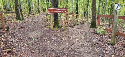 A wooded trail with a sign for "Cut-Through Rock Outdoor Trail" and a directional arrow, surrounded by trees.