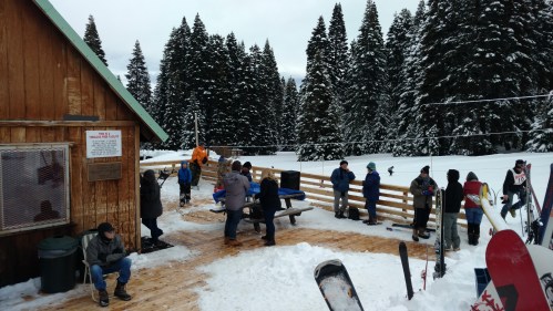 A snowy scene with people gathered outside a wooden lodge, surrounded by tall evergreen trees. Snowboards and skis are present.