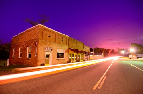 A brick building at dusk with a purple sky, streetlights illuminating a quiet road and light trails from passing cars.