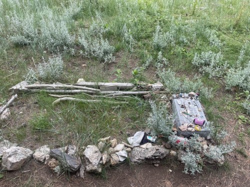 A small, natural memorial site with stones, sticks, and personal items surrounded by grass and plants.