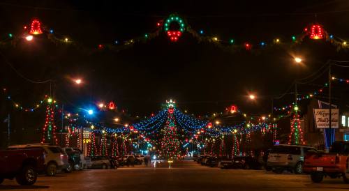 A festive street adorned with colorful holiday lights and decorations, featuring a large illuminated tree at the center.
