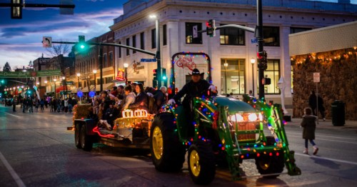A festive tractor pulling a decorated float with people, illuminated by colorful lights, during a parade at dusk.