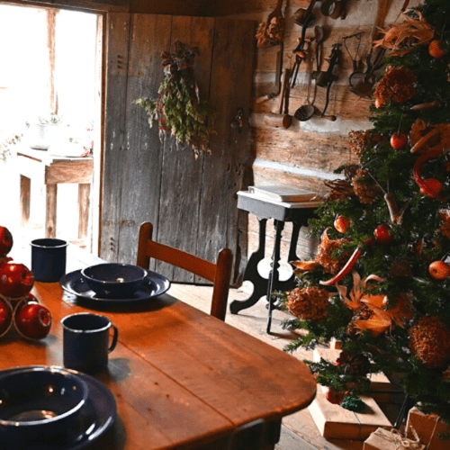 Cozy rustic interior with a wooden table set for a meal, a Christmas tree, and festive decorations.