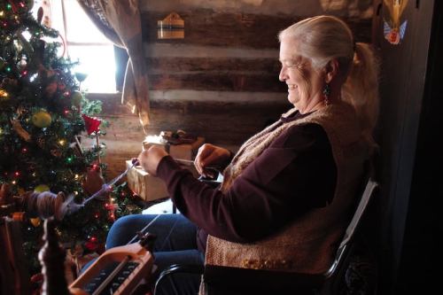A woman with long hair smiles while spinning yarn beside a decorated Christmas tree in a cozy, rustic setting.