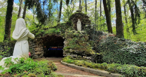 A serene grotto surrounded by trees, featuring a statue of a praying figure and a small shrine.