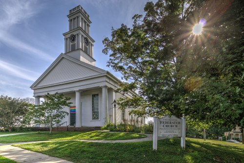 A white church with a clock tower, surrounded by trees, featuring a rainbow flag and a sign that reads "Federated Church."
