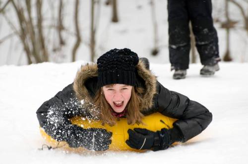 A girl in a black coat and hat joyfully sleds down a snowy hill, with snow flying around her.