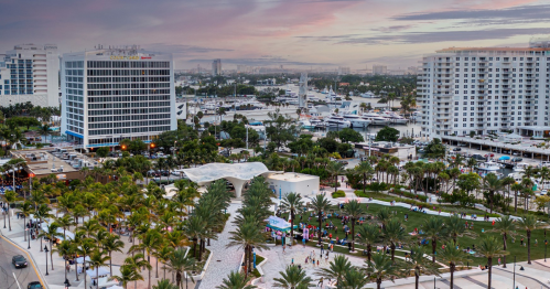 Aerial view of a waterfront cityscape with palm trees, buildings, and boats in the marina under a colorful sky.