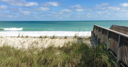 A serene beach scene with gentle waves, sandy shore, and a wooden walkway surrounded by grass under a blue sky.