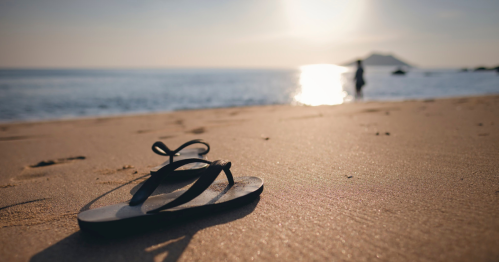 A black flip-flop rests on sandy beach, with a person walking along the shoreline and the sun setting over the water.