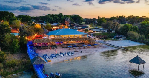 Aerial view of a beachside restaurant at sunset, with lounge chairs, umbrellas, and a calm water shoreline.