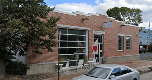 A brick building with large windows, featuring a heart sign, outdoor seating, and a parked car nearby.
