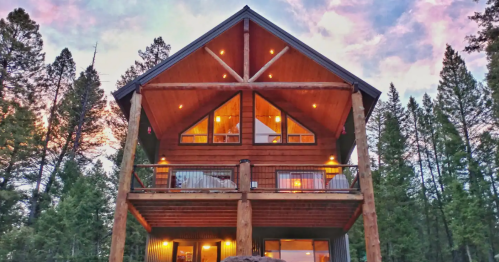 A large wooden cabin with a triangular roof, illuminated windows, and surrounded by trees at sunset.