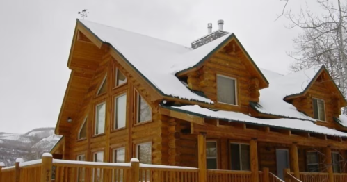 A cozy log cabin with a snow-covered roof, surrounded by trees and mountains in a winter landscape.