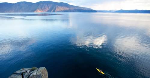 A lone kayaker paddles on a calm lake surrounded by mountains under a partly cloudy sky.