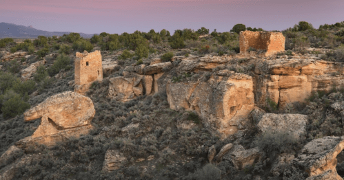 Ancient stone ruins atop rocky terrain, surrounded by greenery and a colorful sky at dusk.