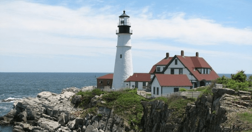 A white lighthouse with a red-roofed house stands on rocky cliffs by the ocean under a clear blue sky.