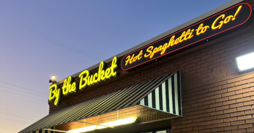 Sign for "By the Bucket Hot Spaghetti to Go!" illuminated on a brick building at dusk.