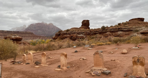 A desert landscape with a historic graveyard, featuring weathered tombstones and rocky formations under a cloudy sky.