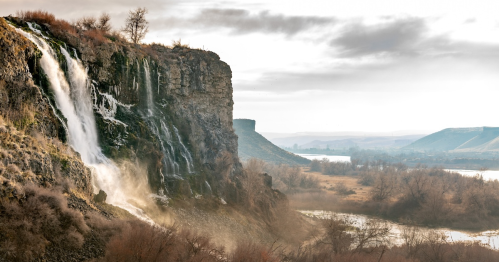 A waterfall cascading down a rocky cliff, surrounded by trees and a river, with a cloudy sky in the background.