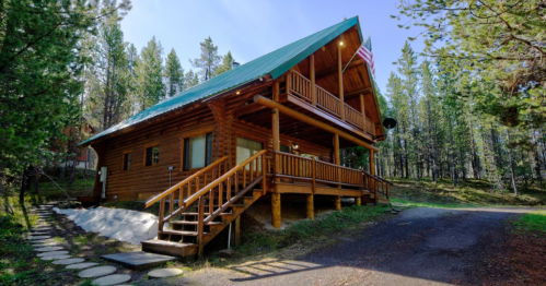 A rustic log cabin with a green roof, surrounded by trees and a gravel path leading up to it.