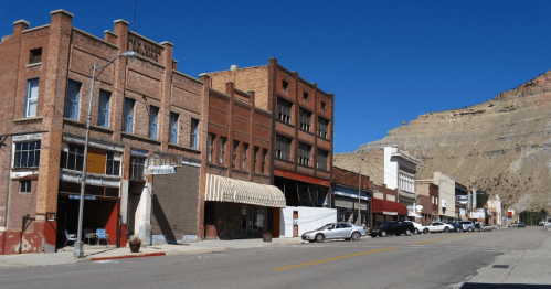A historic street lined with brick buildings and parked cars, set against a clear blue sky and rocky hillside.