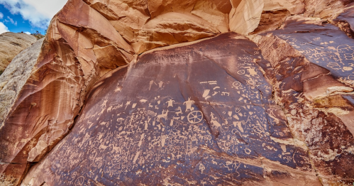 Ancient petroglyphs etched into a rocky cliff, showcasing various symbols and figures against a reddish-brown background.