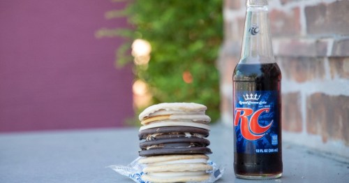 A stack of assorted cookies next to a bottle of RC soda on a table, with a blurred background.